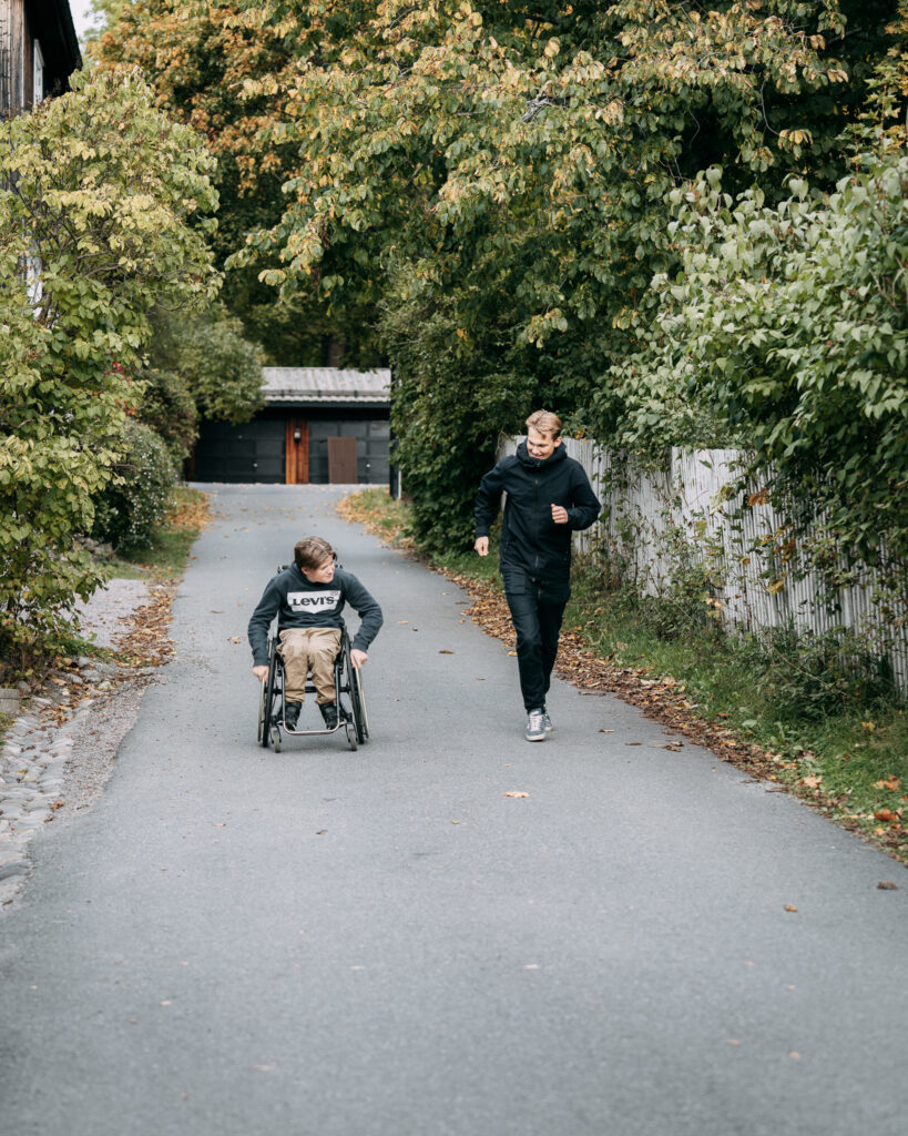 Pojke i rullstol på en väg och manlig personlig assistent springandes bredvid.